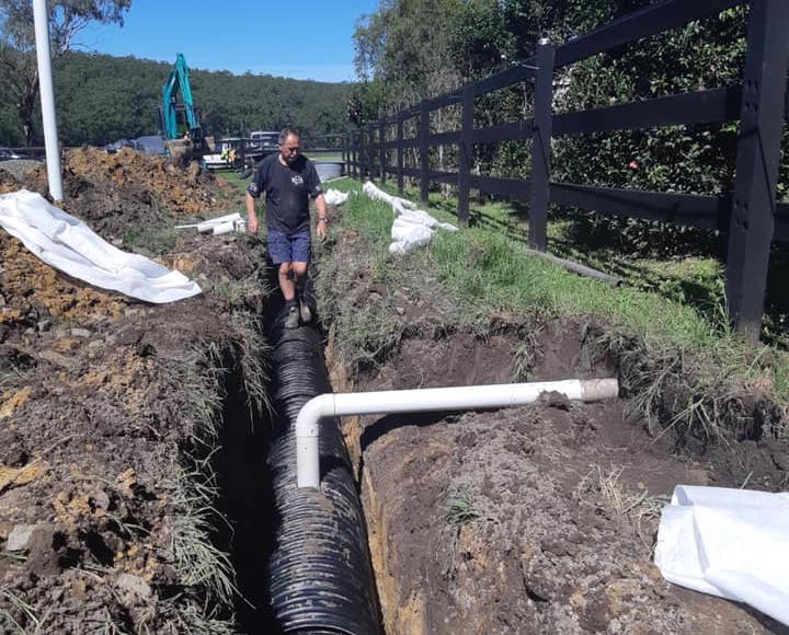 A man standing over a freshly dug trench containing a large black corrugated drainage pipe