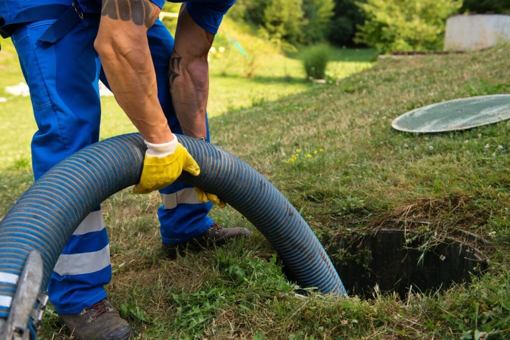 A person in blue workwear and yellow gloves inserts a large blue suction hose into an open underground septic tank