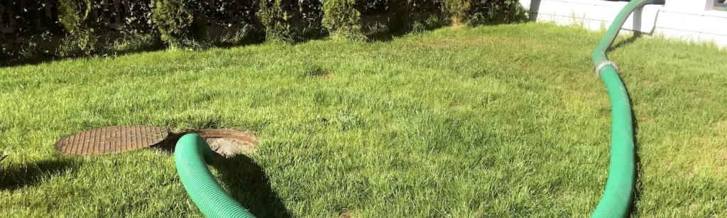 A long green suction hose snakes across a sunny green lawn, with one end disappearing into an open manhole.