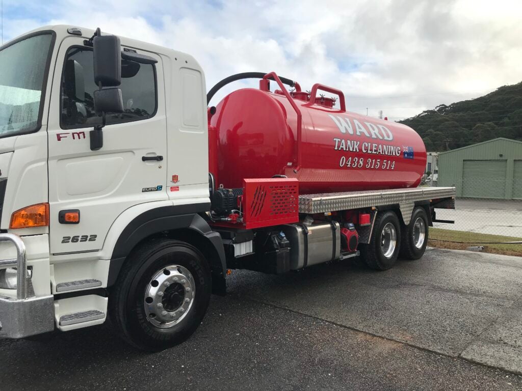 A front-angled view of a white Ward Tank Cleaning truck with a red vacuum tank, parked on a wet asphalt surface.