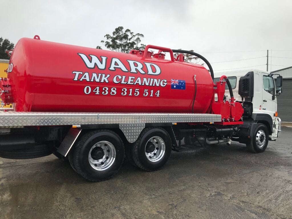 A white service truck with a large red vacuum tank labelled "WARD TANK CLEANING" and a phone number, parked on a wet paved surface.