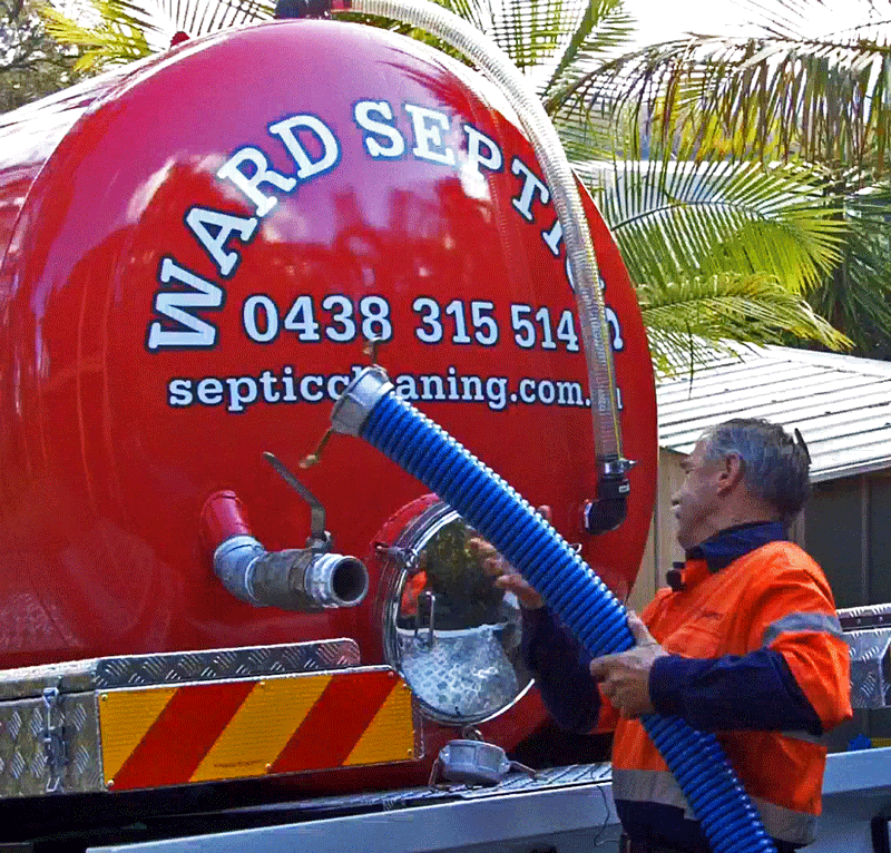A technician in a high-visibility orange shirt connecting a large blue suction hose to the rear of a red "WARD SEPTIC" vacuum tank.
