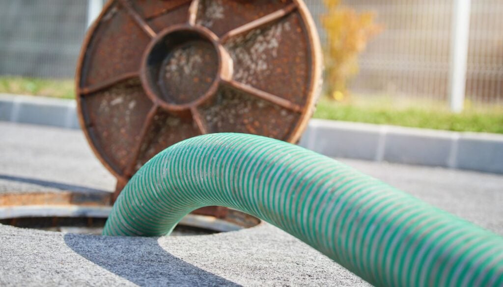 A close-up of a thick green suction hose inserted into an open manhole on an asphalt surface.