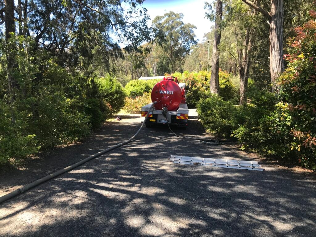 Ward Tank Cleaning truck parked on a shaded gravel driveway, with a long suction hose extending out toward a property.