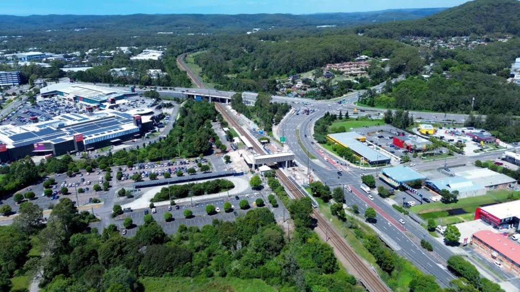 A busy suburban interchange featuring a train station on a railway line that passes under a multi-lane road, with a large shopping center and parking lots surrounded by lush green trees and hills.