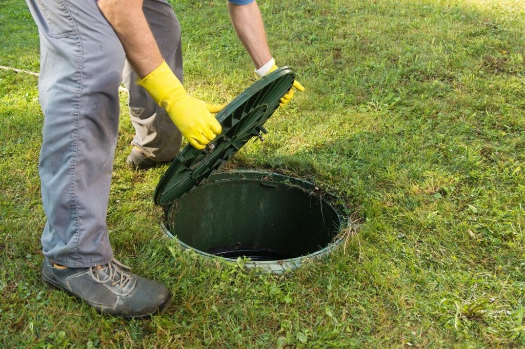 A person wearing yellow gloves opens a green circular lid of an underground septic tank or utility access point in a grassy field.