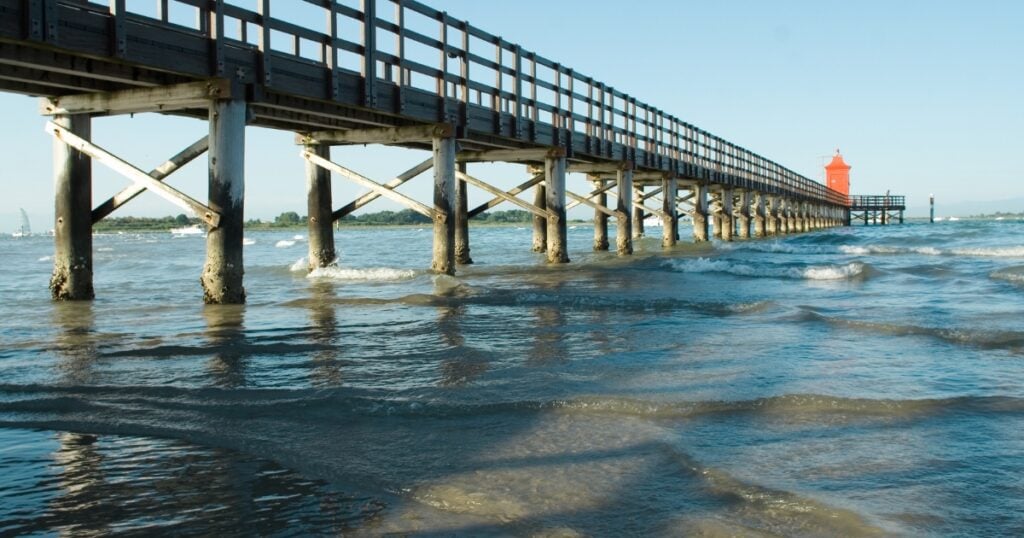 A long wooden pier with cross-braced pilings stretching over choppy ocean water toward a small red lighthouse structure at the far end under a clear blue sky.