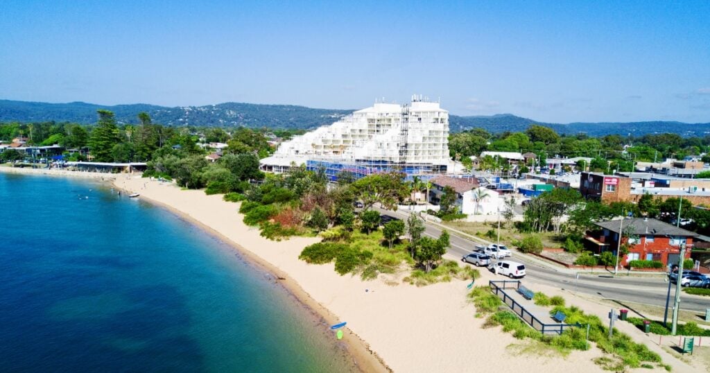 A sandy beach curving along turquoise ocean water, featuring a large white stepped resort hotel building.