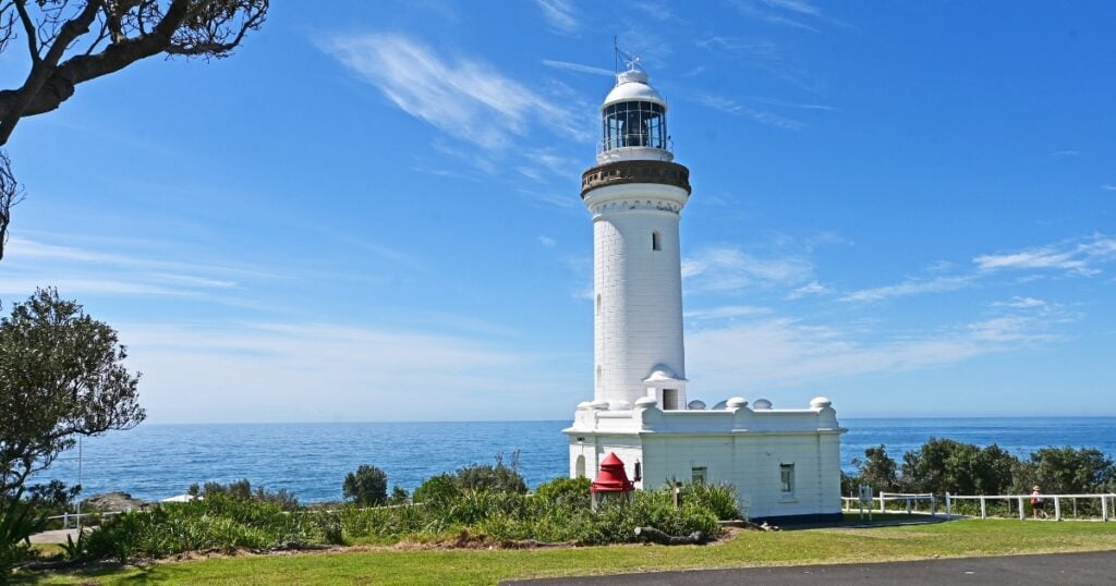 A white lighthouse on a grassy hill overlooking the blue ocean under a partly cloudy sky in Norah Head