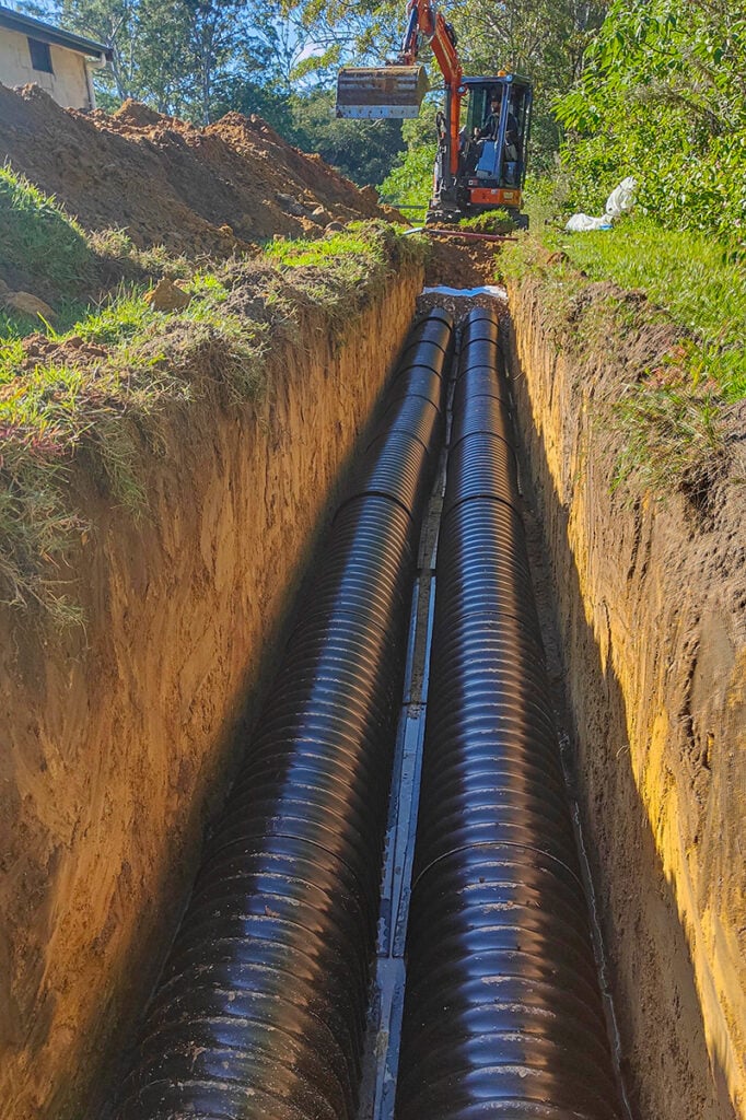 A long, deep dirt trench looking up toward a small excavator, with two large, black ribbed drainage pipes.