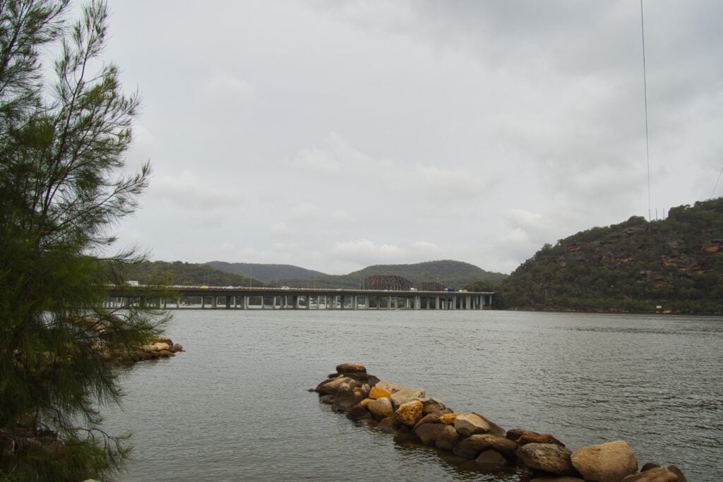 A wide bridge spans a river under an overcast sky, with forested hills in the background and a rocky shoreline in the foreground.