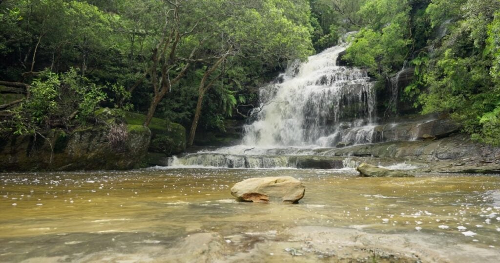 A beautiful multi-tiered waterfall cascading over dark rocks into a shallow, sandy-bottomed pool in Somersby.