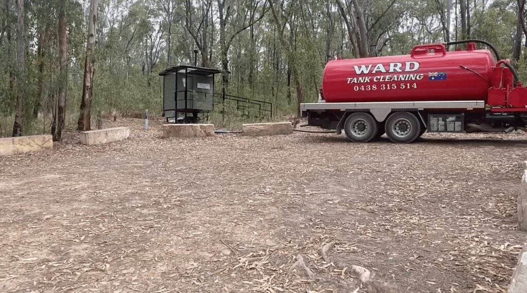 A red Ward Tank Cleaning truck is parked in a wooded clearing next to a small corrugated metal outhouse or utility shed.