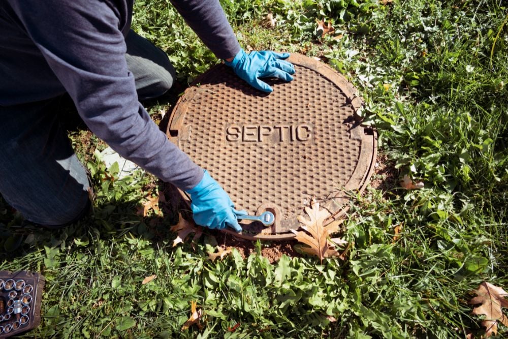 A worker wearing blue latex gloves uses a socket wrench to secure or loosen a heavy metal septic tank cover labelled "SEPTIC" in a grassy lawn.