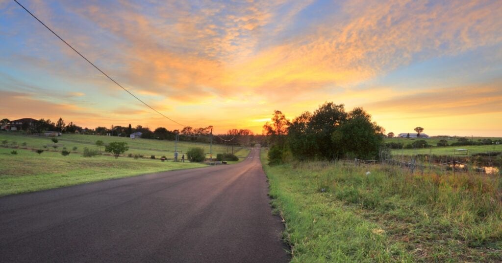 A two-lane asphalt road stretching through rural rolling green hills and farmland in Bucketty