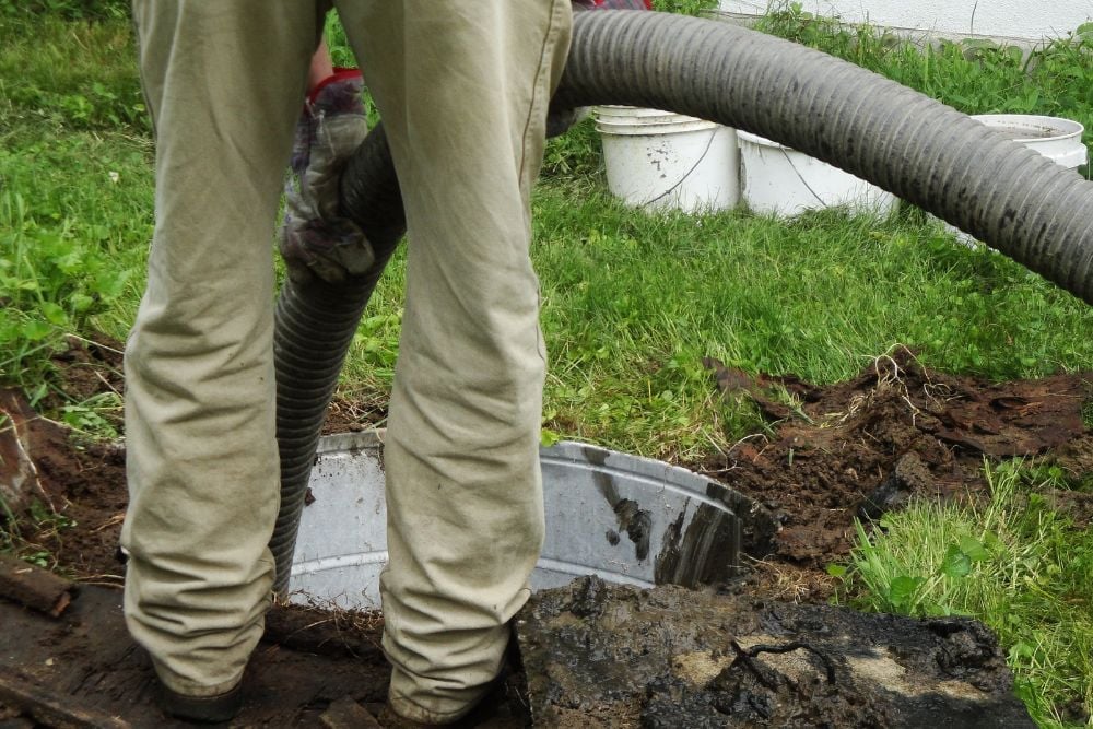 A worker in tan trousers and work gloves guiding a large, grey ribbed suction hose into an open concrete septic tank riser