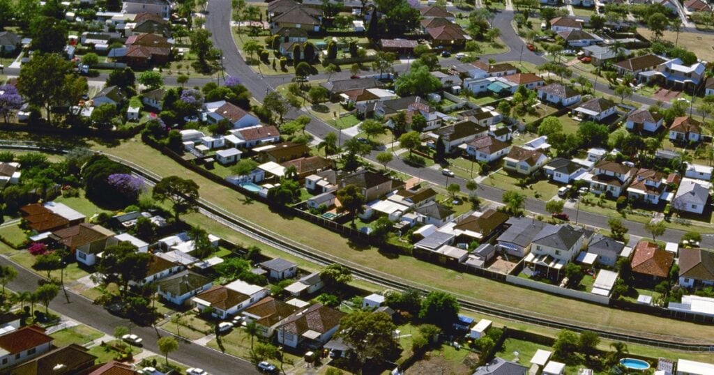 A densely populated suburban neighborhood with closely spaced single-storey homes, green lawns, paved streets, and a long grassy easement in Laguna.