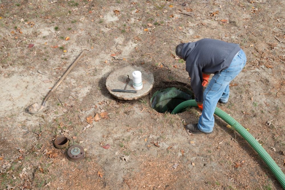 A worker in a grey hoodie and blue jeans inserts a thick green suction hose into an open underground septic tank