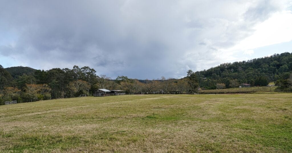 An expansive, grassy field in a rural countryside landscape featuring a small rustic barn or shed.