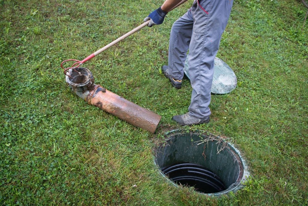 A worker using a long-handled hook tool to pull a large, dirty plastic pipe assembly out of an open septic tank riser in a grassy yard.