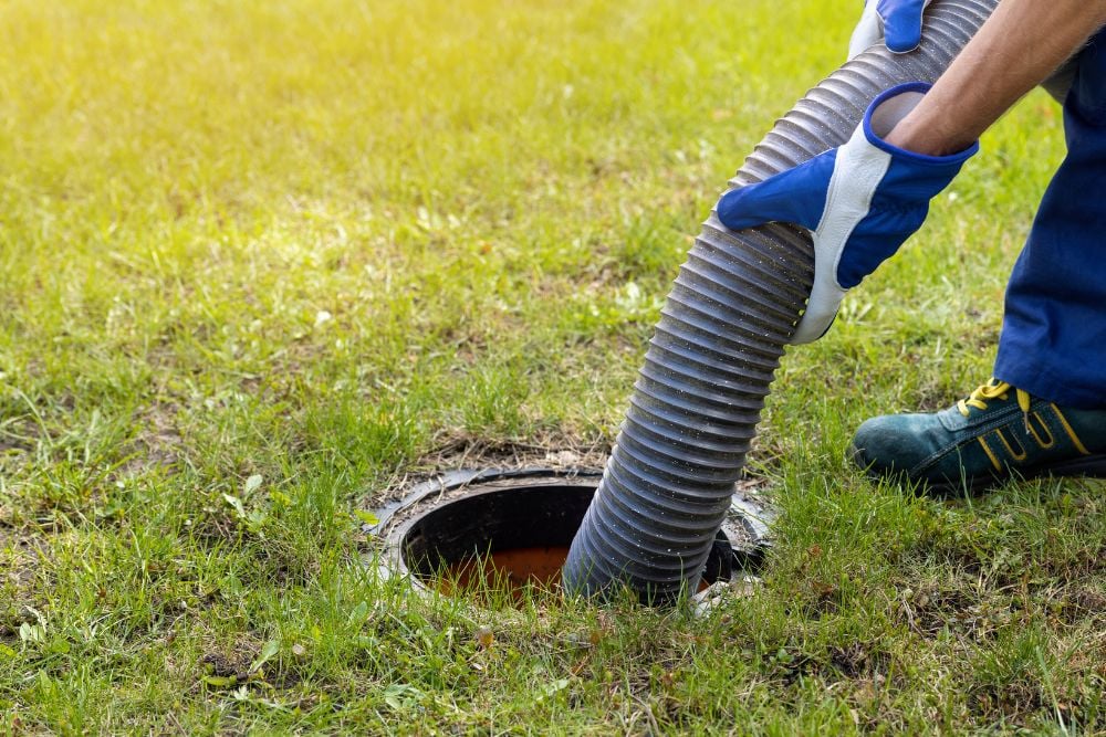 A close-up of a worker in blue and white gloves guiding a ribbed grey suction hose into an open septic tank hole in a green lawn.