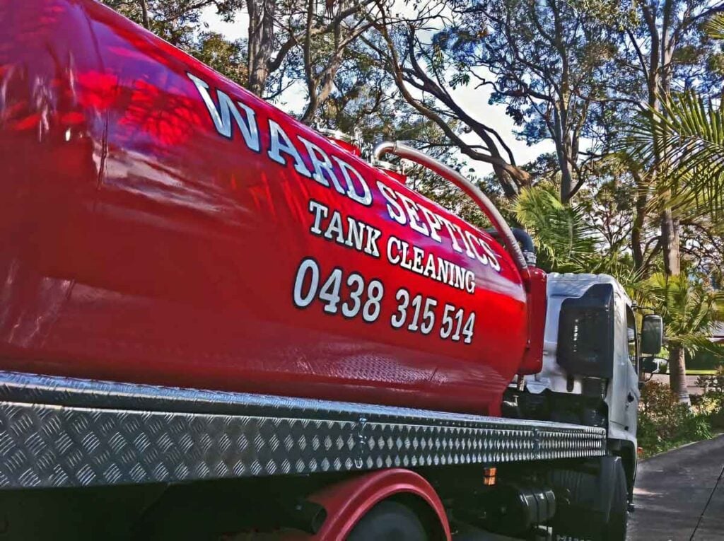 A shiny red Ward Septics tank cleaning truck is parked on a driveway lined with trees.