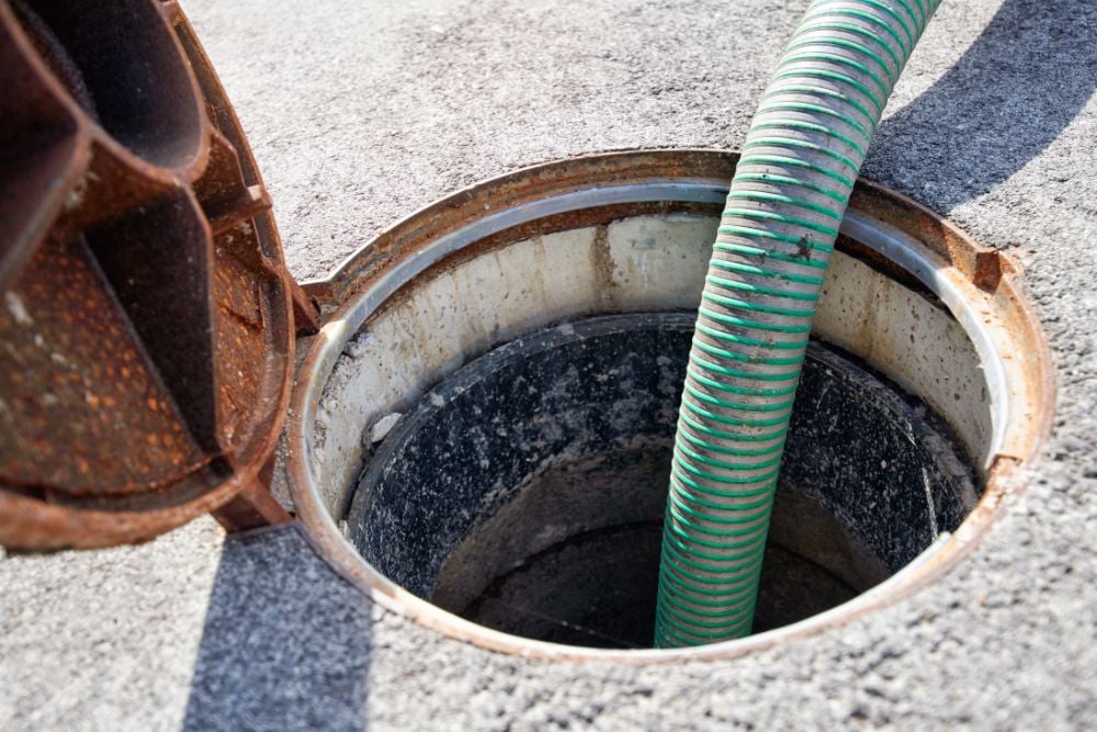 A green ribbed suction hose inserted into an open, circular manhole on an asphalt surface.