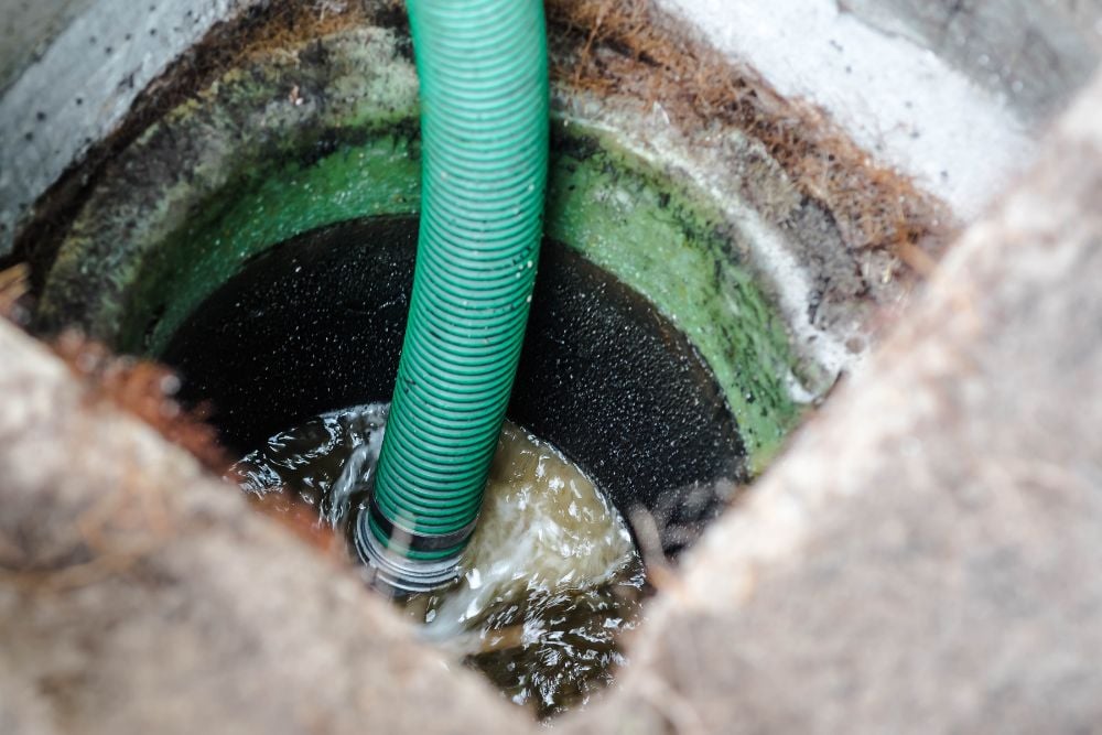 A green suction hose submerged in churning, murky water inside a concrete septic tank riser during a pumping process.