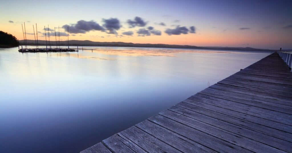A long wooden pier extends diagonally over calm, glassy water at sunset in Tuggerah Lake.