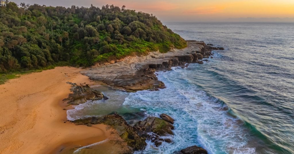 An elevated view of ocean waves crashing against a rocky headland and sandy beach in Wamberal Lagoon.