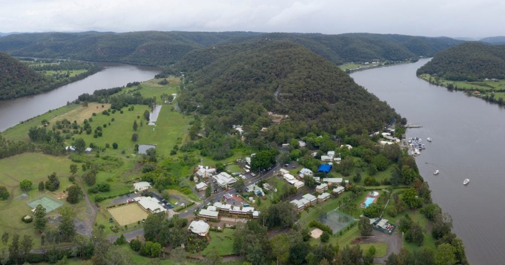 A small riverside town nestled along a wide bend in a river, bordered by rolling, densely forested hills in Wisemans Ferry