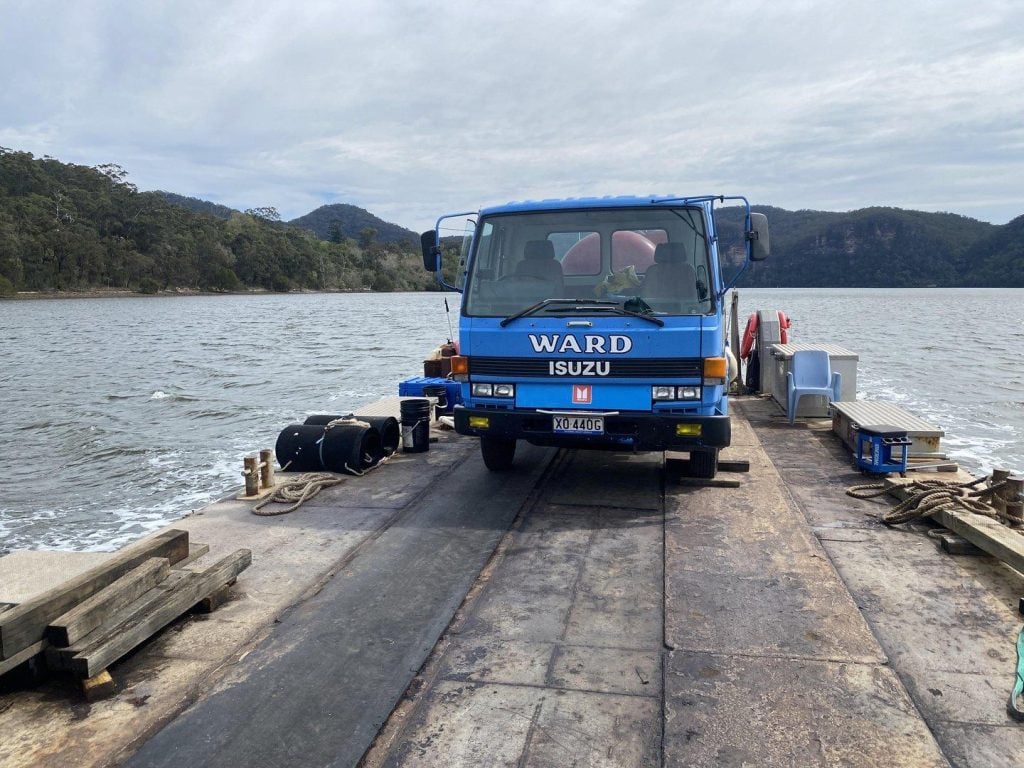 A blue Isuzu truck with "WARD" branding parked on a flat barge or ferry traveling across a river.