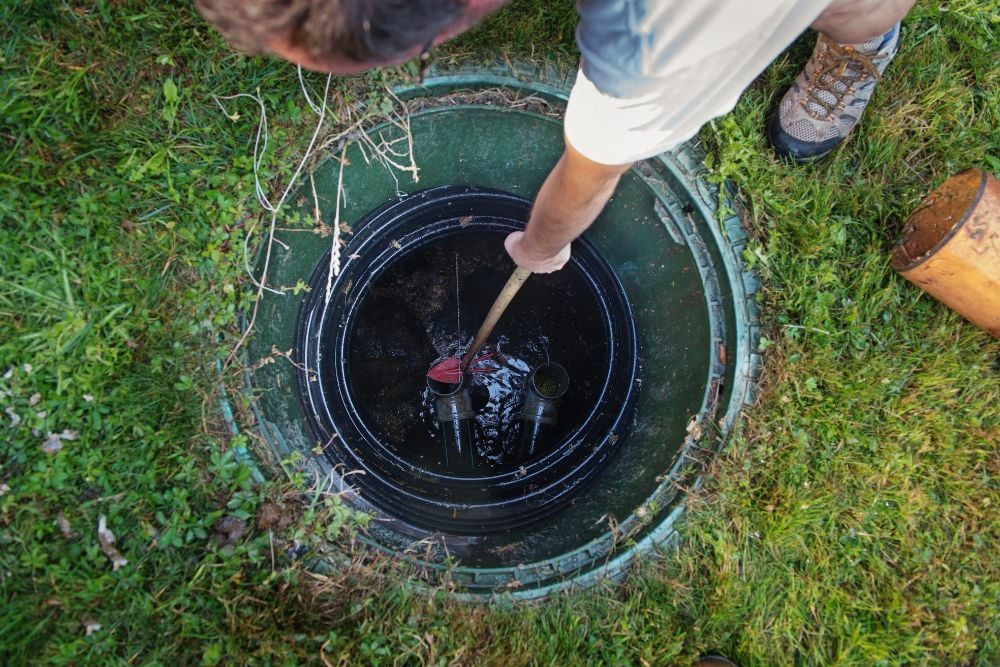 A top-down view of a worker using a long wooden tool to inspect or clear debris within an open green septic tank riser