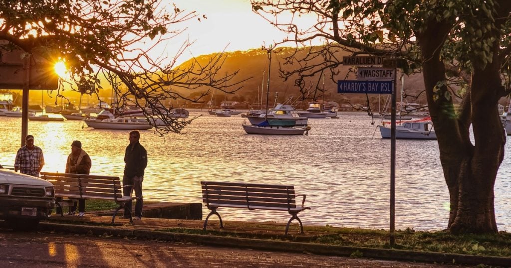 A waterfront path with park benches at sunset, looking out over a bay filled with anchored boats and directional road signs in Wagstaffe.