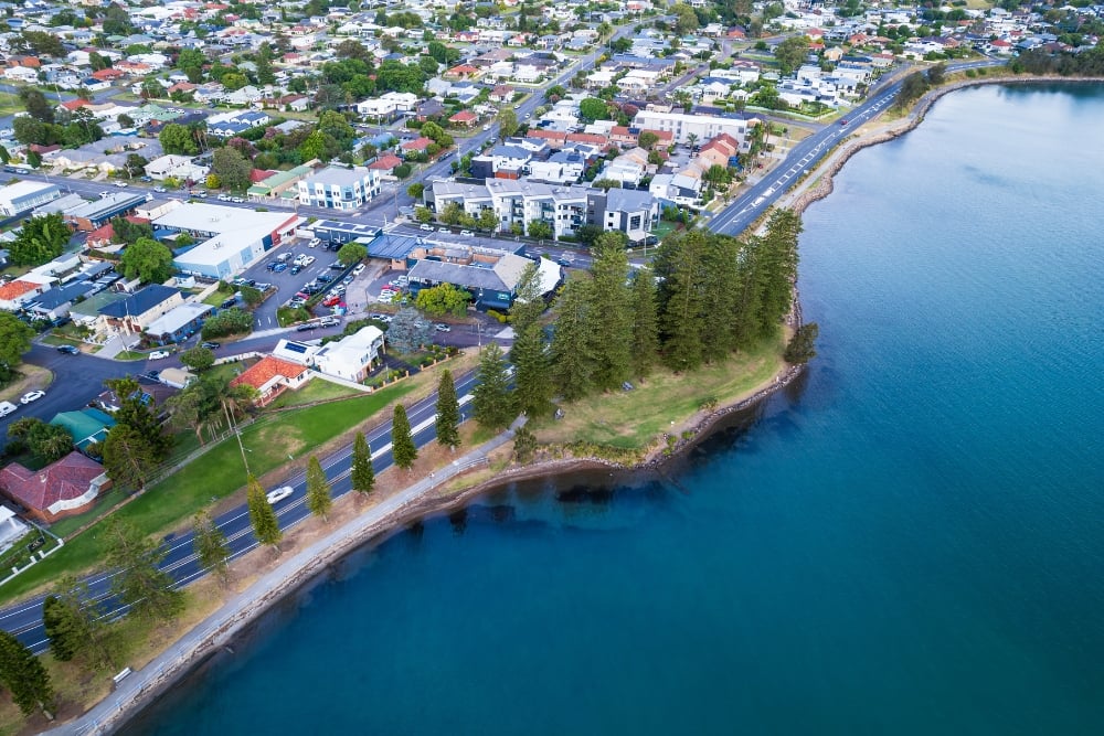 A coastal suburban town bordered by a clear blue body of water, with a tree-lined main road running parallel to the shoreline in Bonnells Bay
