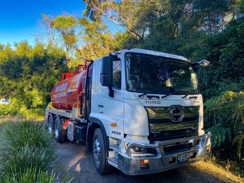 A white Hino Ward Tank Cleaning truck parked on a gravel path surrounded by lush trees.